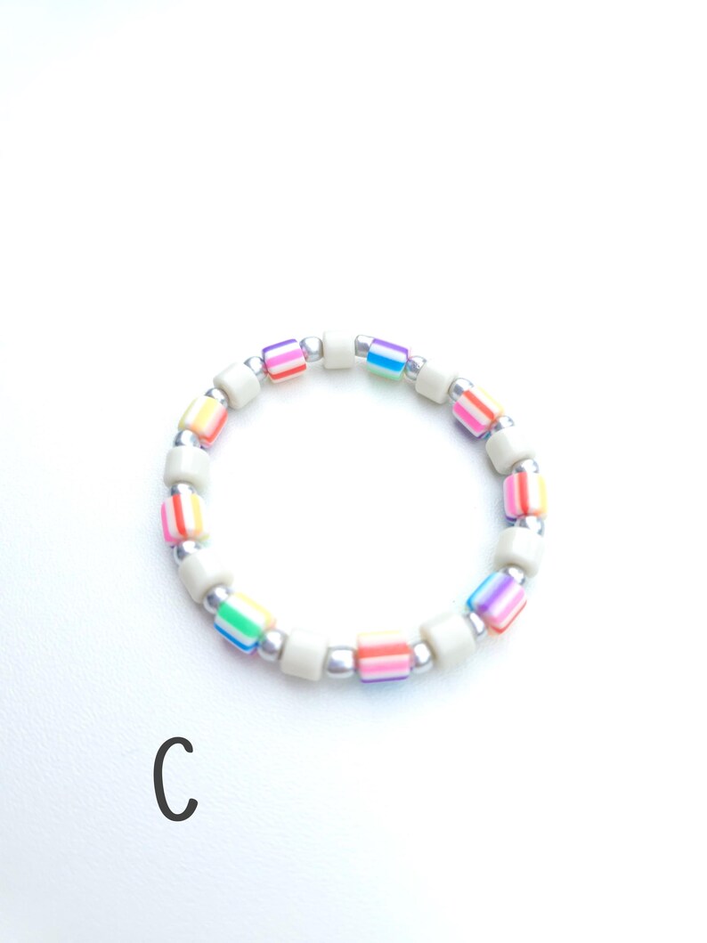 May include: A circular beaded bracelet featuring alternating segments of solid off-white cylindrical beads, metallic silver spherical beads, and multi-colored striped cylindrical beads in shades of pink, orange, yellow, green, blue, and purple. The letter C is visible.