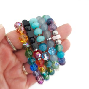 K&ouml;nnte beinhalten: Eine Sammlung von f&uuml;nf gestapelten Armb&auml;ndern mit einer Vielzahl von Perlen, darunter facettiertes Glas, glatte runde Steine und schwarze quadratische W&uuml;rfel. Die Perlen zeigen ein Farbspektrum wie Rot, Orange, Gelb, Gr&uuml;n, Blau und Violett.