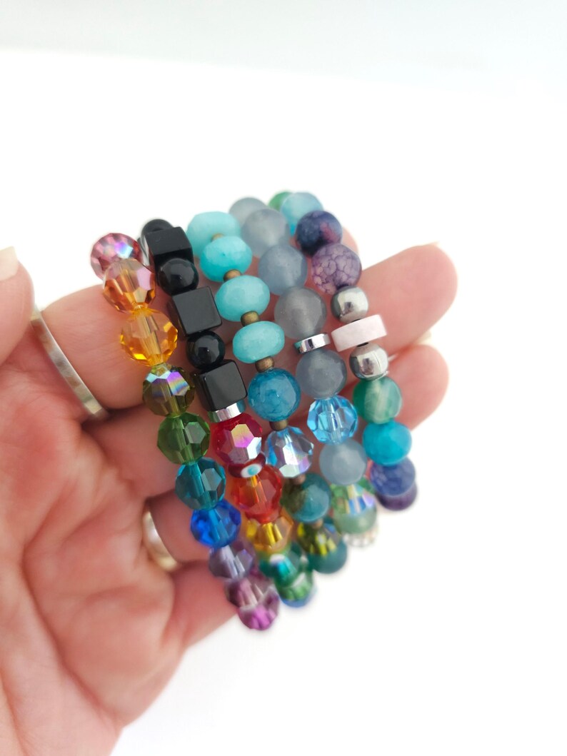 K&ouml;nnte beinhalten: Eine Sammlung von f&uuml;nf gestapelten Armb&auml;ndern mit einer Vielzahl von Perlen, darunter facettiertes Glas, glatte runde Steine und schwarze quadratische W&uuml;rfel. Die Perlen zeigen ein Farbspektrum wie Rot, Orange, Gelb, Gr&uuml;n, Blau und Violett.