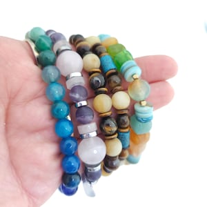 May include: A collection of four beaded bracelets featuring various natural stones in shades of blue, purple, pink, brown, and green. The bracelets are constructed with round and disc-shaped beads, interspersed with metallic silver and gold-toned spacers.