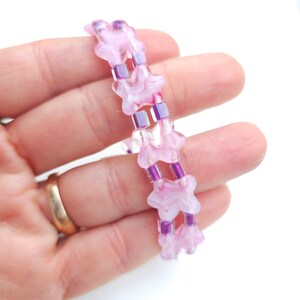 May include: A beaded bracelet featuring two parallel strands of light pink, translucent, flower-shaped beads alternating with small, rectangular, metallic purple spacer beads. The design creates a repeating pattern of floral and geometric elements.