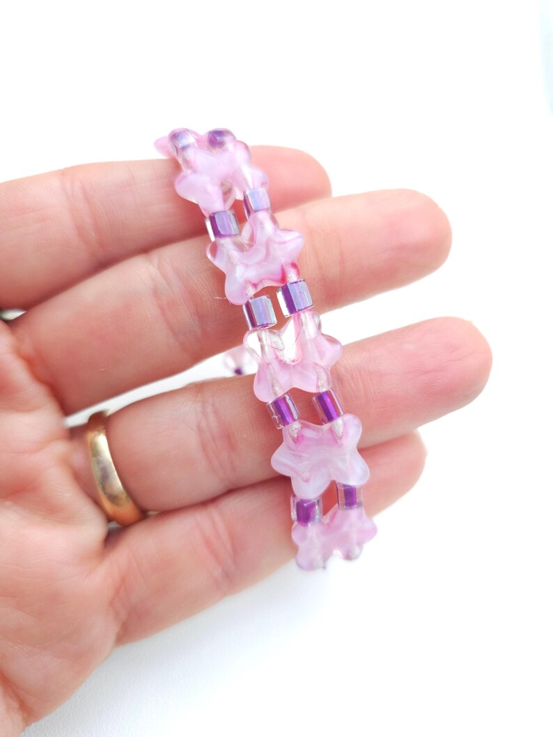 May include: A beaded bracelet featuring two parallel strands of light pink, translucent, flower-shaped beads alternating with small, rectangular, metallic purple spacer beads. The design creates a repeating pattern of floral and geometric elements.