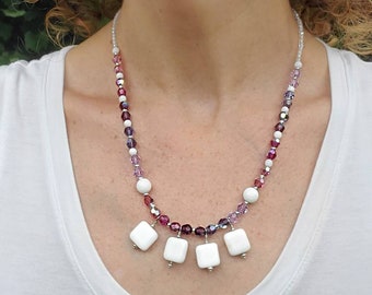 May include: A beaded necklace featuring white, purple, and clear beads. The necklace has a gradient of colors, with white beads and square pendants at the bottom. The necklace is worn against a white shirt.