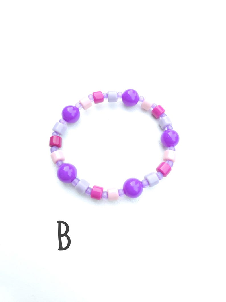 May include: A circular bracelet featuring a repeating pattern of purple spherical beads, light purple cylindrical beads, pink square beads, and light pink cylindrical beads strung together. The letter B is printed in black below the bracelet.