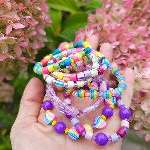 May include: A collection of stacked bracelets made from colorful plastic beads in various shapes and sizes, including round, square, and heart-shaped beads in shades of purple, yellow, pink, blue, green, and orange, featuring striped and marbled patterns.