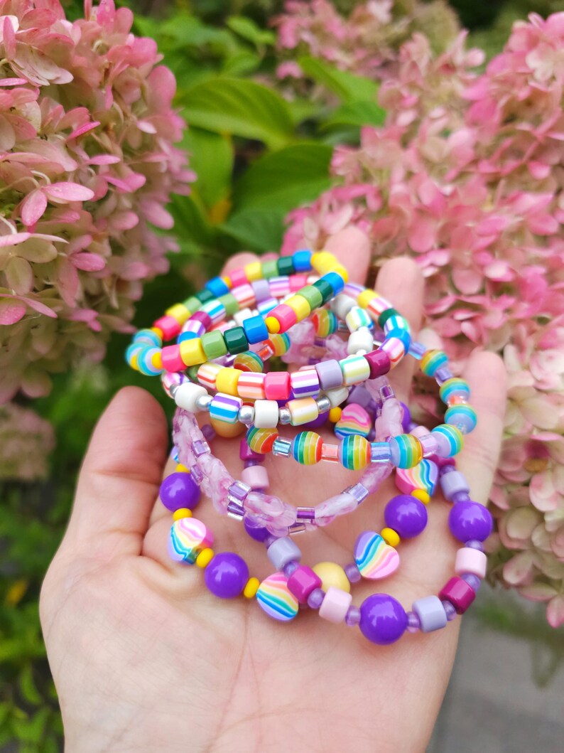 May include: A collection of stacked bracelets made from colorful plastic beads in various shapes and sizes, including round, square, and heart-shaped beads in shades of purple, yellow, pink, blue, green, and orange, featuring striped and marbled patterns.