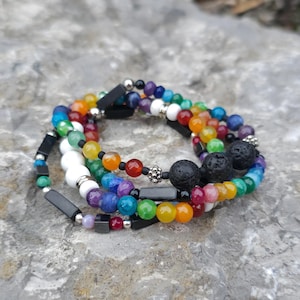 May include: A stack of beaded bracelets in a variety of colors, including red, orange, yellow, green, blue, and purple. The bracelets feature a mix of round and rectangular beads, with some accented by silver-colored beads and black rectangular beads. The bracelets are stacked on a gray stone surface.