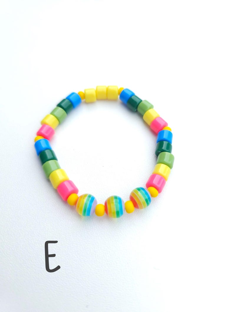 May include: A circular bracelet featuring a repeating pattern of cylindrical beads in yellow, pink, blue, and green, interspersed with small yellow spacer beads and three larger spherical beads with multicolored vertical stripes, marked with the letter E.