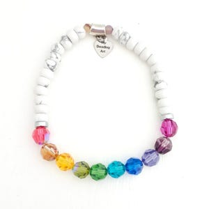 K&ouml;nnte beinhalten: Ein elastisches Armband mit wei&szlig;en marmorierten Perlen und einer Reihe facettierter regenbogenfarbener Perlen. Ein silberfarbener herzf&ouml;rmiger Anh&auml;nger h&auml;ngt oben und ist mit dem Text My Beading Art graviert. Die Perlen sind kreisf&ouml;rmig angeordnet.