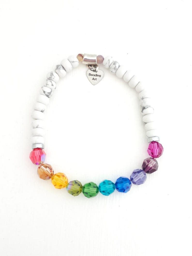 K&ouml;nnte beinhalten: Ein elastisches Armband mit wei&szlig;en marmorierten Perlen und einer Reihe facettierter regenbogenfarbener Perlen. Ein silberfarbener herzf&ouml;rmiger Anh&auml;nger h&auml;ngt oben und ist mit dem Text My Beading Art graviert. Die Perlen sind kreisf&ouml;rmig angeordnet.