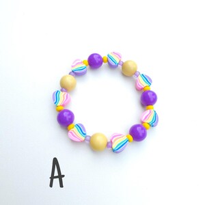 May include: A circular bracelet featuring alternating purple spheres, yellow spheres, and heart-shaped beads with rainbow stripes. The beads are strung together with small clear spacers. The letter A is printed in black below the bracelet.