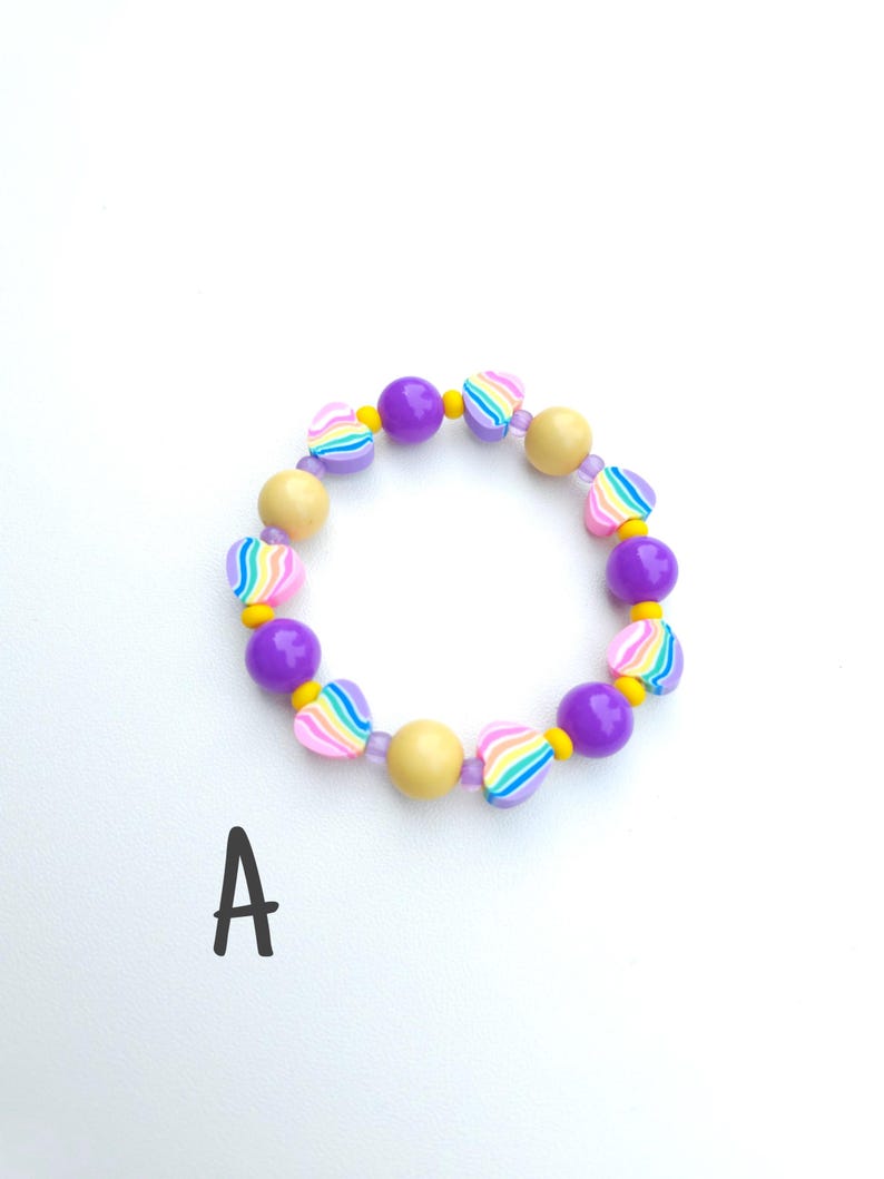 May include: A circular bracelet featuring alternating purple spheres, yellow spheres, and heart-shaped beads with rainbow stripes. The beads are strung together with small clear spacers. The letter A is printed in black below the bracelet.