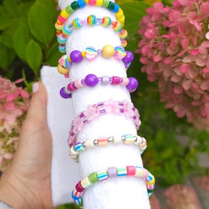 May include: A collection of seven colorful beaded bracelets stacked on a white cylindrical display. The bracelets feature a variety of plastic beads in rainbow colors, including striped, solid, translucent, and flower-shaped designs.