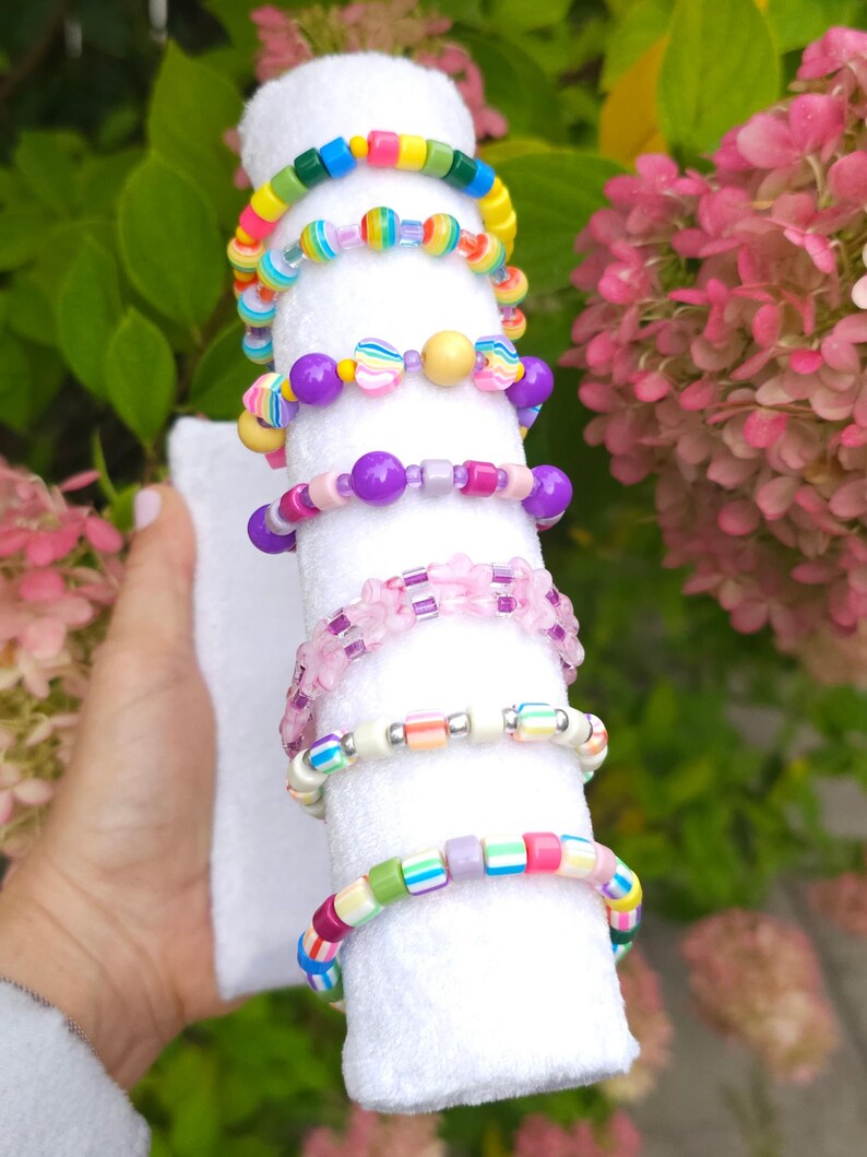 May include: A collection of seven colorful beaded bracelets stacked on a white cylindrical display. The bracelets feature a variety of plastic beads in rainbow colors, including striped, solid, translucent, and flower-shaped designs.