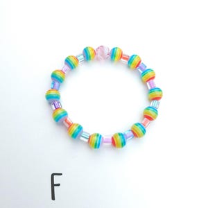 May include: A circular beaded bracelet featuring alternating round beads with horizontal rainbow stripes and small translucent square spacer beads. A single light pink faceted bead is positioned at the top. The letter F is printed below.