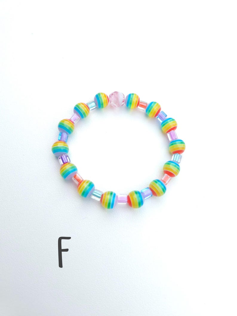 May include: A circular beaded bracelet featuring alternating round beads with horizontal rainbow stripes and small translucent square spacer beads. A single light pink faceted bead is positioned at the top. The letter F is printed below.