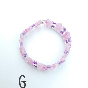 May include: A circular beaded bracelet featuring alternating light pink marbled star-shaped beads and small translucent purple cylindrical beads. The bracelet is secured with a single silver-toned metallic bead. The letter G is printed in black.
