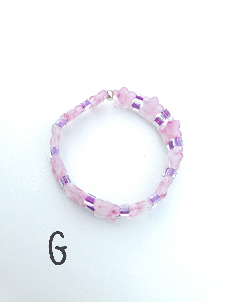 May include: A circular beaded bracelet featuring alternating light pink marbled star-shaped beads and small translucent purple cylindrical beads. The bracelet is secured with a single silver-toned metallic bead. The letter G is printed in black.