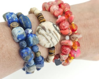 May include: A set of three beaded bracelets. One features blue and grey stone nuggets with brass spacers. The second has small tan beads, brown disc spacers, and a large round patterned focal bead. The third consists of red and orange stone beads.