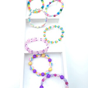 May include: A collection of six colorful beaded bracelets arranged in a white tiered display. The bracelets feature various bead shapes including round, square, and star-shaped beads in vibrant hues of pink, purple, yellow, blue, and green.