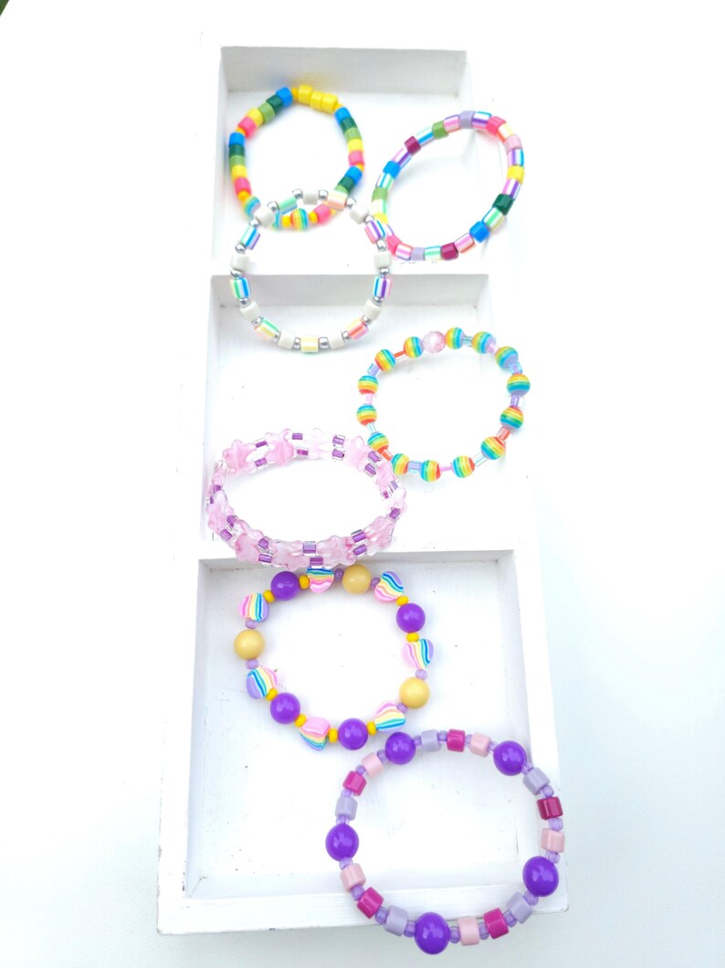 May include: A collection of six colorful beaded bracelets arranged in a white tiered display. The bracelets feature various bead shapes including round, square, and star-shaped beads in vibrant hues of pink, purple, yellow, blue, and green.