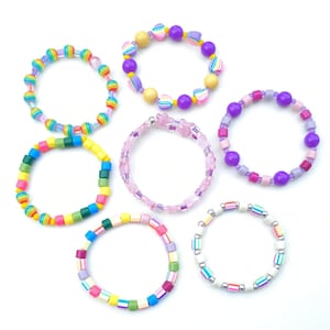 May include: A collection of six circular beaded bracelets featuring various designs. One bracelet has rainbow-striped beads, another features yellow and purple spheres with heart accents, and one is made of translucent pink star-shaped beads. Additional bracelets include multi-colored square beads, purple and pink spheres, and white beads with colorful stripes.