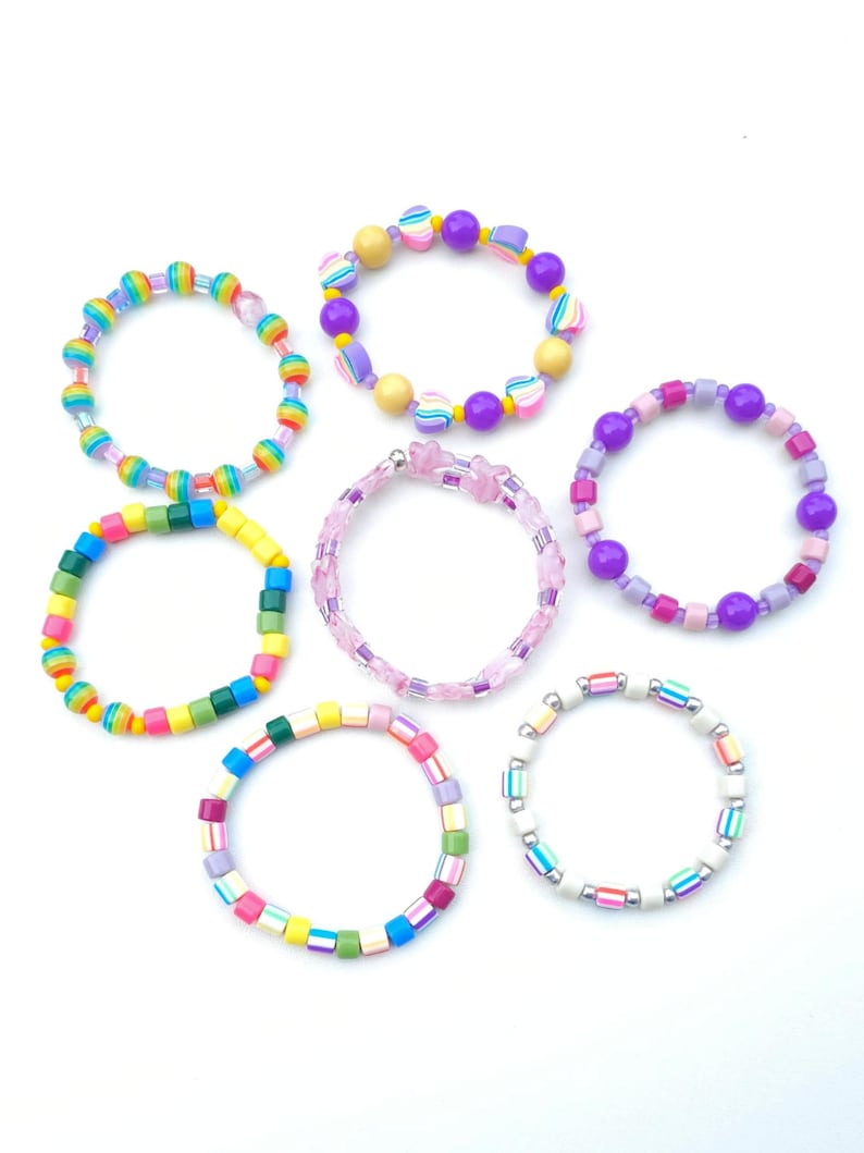 May include: A collection of six circular beaded bracelets featuring various designs. One bracelet has rainbow-striped beads, another features yellow and purple spheres with heart accents, and one is made of translucent pink star-shaped beads. Additional bracelets include multi-colored square beads, purple and pink spheres, and white beads with colorful stripes.