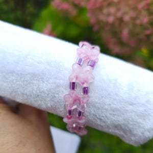 May include: A beaded bracelet featuring alternating translucent pink flower-shaped beads and small square purple beads. The bracelet is constructed with a repeating pattern of one flower bead followed by two purple square beads in a linear design.