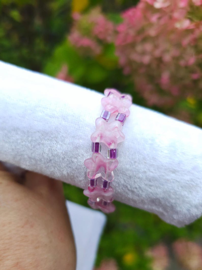 May include: A beaded bracelet featuring alternating translucent pink flower-shaped beads and small square purple beads. The bracelet is constructed with a repeating pattern of one flower bead followed by two purple square beads in a linear design.