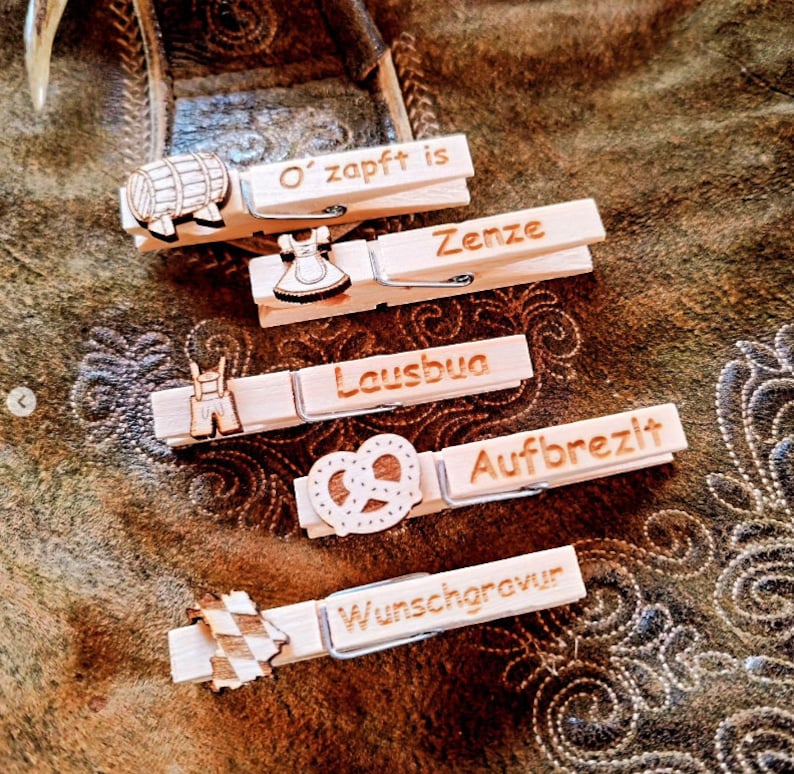 May include: Five wooden clothespins with different designs and text. The clothespins are decorated with a beer barrel, a dirndl, lederhosen, a pretzel, and a Bavarian flag. The text on the clothespins reads "O' zapft is", "Zenze", "Lausbua", "Aufbrezlt", and "Wunschgravur".