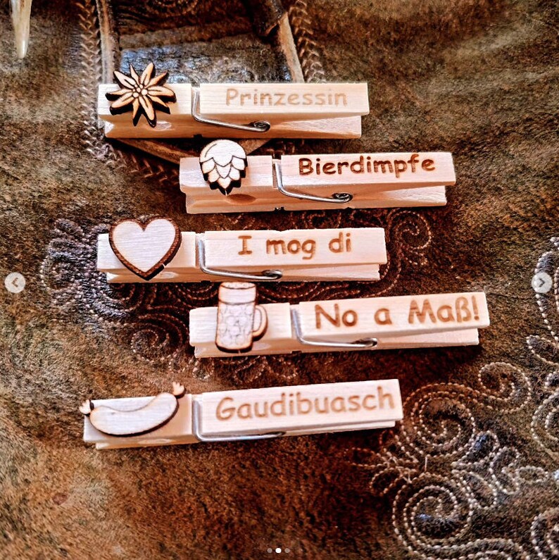 May include: Five wooden clothespins with different designs and text. The designs include a heart, a beer mug, a pretzel, a sausage, and an edelweiss flower. The text on the clothespins reads "Prinzessin", "Bierdim pfe", "I mog di", "No a Ma&szlig;!" and "Gaudibuasch".