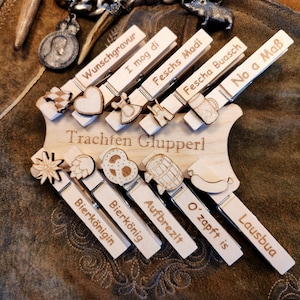 Könnte beinhalten: Holz-Wäscheklammern mit verschiedenen deutschen Sprüchen und Bildern, darunter ein Brezel, ein Bierkrug, ein Herz und eine Wurst. Die Wäscheklammern sind auf einem Holzbrett mit dem Text "Trachten Glupperl" angeordnet.