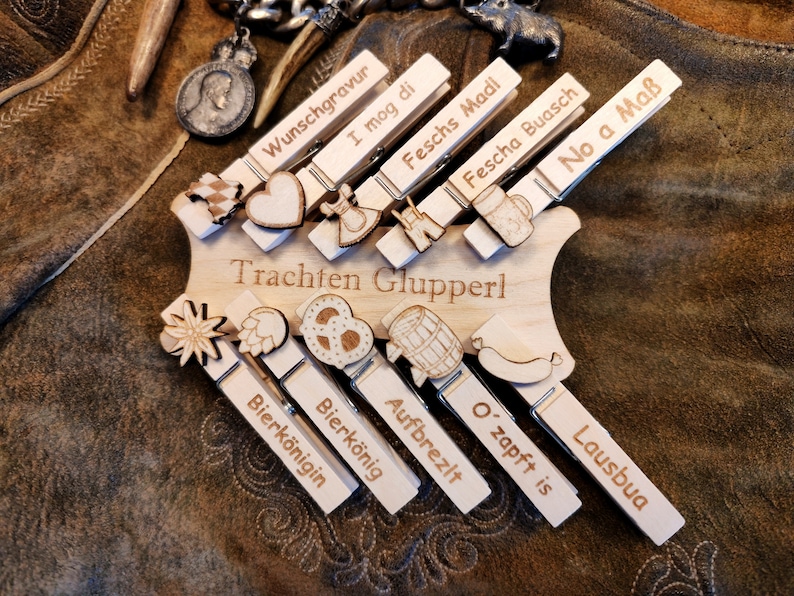 May include: Wooden clothespins with various German phrases and images, including a pretzel, a beer stein, a heart, and a sausage. The clothespins are arranged on a wooden board with the text "Trachten Glupperl".