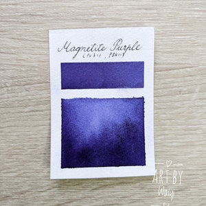 May include: A watercolor paint swatch card showing the color Magnetite Purple (PBk7) with a description of the color as "C.I. PBk7". The card is on a light wood surface. The card has the text "ART BY Waivy" with a heart on each side.