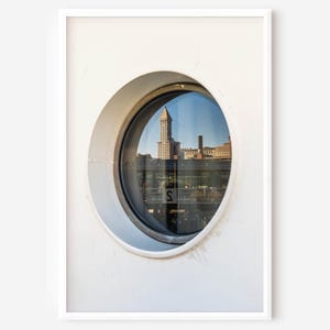 May include: A framed photograph featuring a round window with a city view reflected in its glass. The window is set in a white wall. The reflection shows a tall building and other structures, with the letter "S" visible.