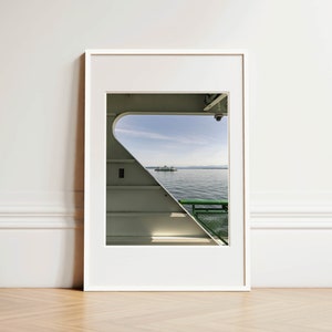 A vertical photograph of a Washington State Ferry boat gliding through Puget Sound, framed in the image by the unique angled architecture of another ferry boat.  Mountains and blue sky in background, green railing and ferry architecture in foreground