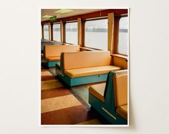 May include: A photograph of a ferry interior featuring rows of orange vinyl bench seats with teal bases arranged along large rectangular windows. The floor has alternating brown and tan stripes, and a green sign reading RESTROOM is visible.