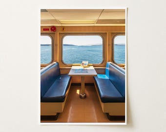 May include: A ferry seating area featuring two blue vinyl bench seats facing each other with a central white table. Large rectangular windows provide a clear view of the blue ocean and distant horizon under a bright sky. A red circular light is visible.