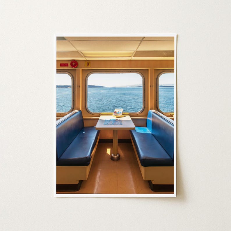May include: A ferry seating area featuring two blue vinyl bench seats facing each other with a central white table. Large rectangular windows provide a clear view of the blue ocean and distant horizon under a bright sky. A red circular light is visible.