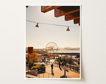 May include: A photograph of an outdoor waterfront promenade featuring a large Ferris wheel in the distance under a clear sky. The scene includes wooden overhead beams, hanging string lights, paved walkways, and people walking near trees.