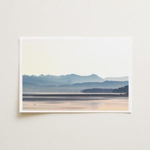 May include: A rectangular landscape print featuring a serene lake reflecting soft light, with layers of blue-toned mountain silhouettes in the distance and a dark, forested shoreline on the right side under a pale, bright sky.