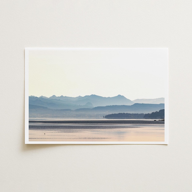 May include: A rectangular landscape print featuring a serene lake reflecting soft light, with layers of blue-toned mountain silhouettes in the distance and a dark, forested shoreline on the right side under a pale, bright sky.