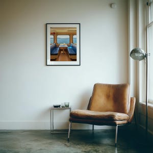 May include: A framed wall art print depicting a ferry interior with blue bench seating, a small table, and large windows overlooking the ocean. The print is displayed in a black frame on a white wall above a brown leather armchair and a metal side table.