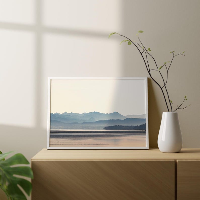 May include: A framed landscape print featuring a serene lake reflecting a pale sky with layered blue mountain silhouettes in the distance. The artwork is displayed in a thin white frame, positioned next to a white vase containing a thin branch.