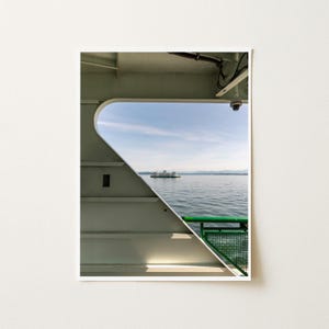 May include: A view from inside a ferry deck looking out through a large triangular window at a distant ferry on the water. The interior structure is off-white with a green metal railing visible in the lower right corner of the window frame.