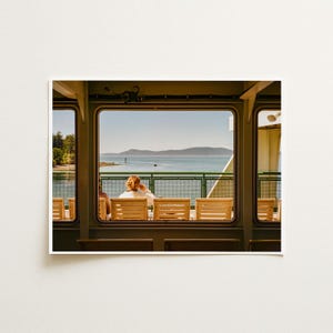May include: A view through a rectangular ferry window showing rows of light-colored wooden chairs facing a green metal railing. Beyond the railing, a calm body of water stretches toward distant hills under a clear, bright blue sky.