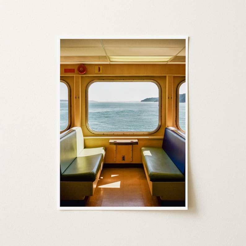 May include: A view from inside a ferry cabin featuring two facing bench seats with green cushions and blue backrests. A large rectangular window centered between the seats looks out over a calm body of water toward a distant shoreline.
