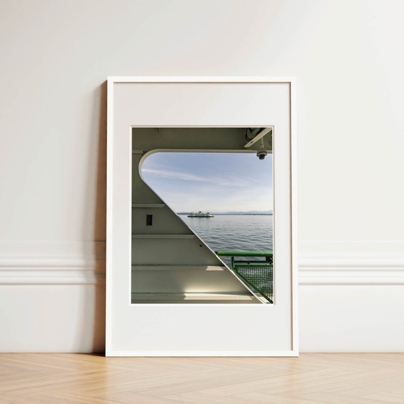 A vertical photograph of a Washington State Ferry boat gliding through Puget Sound, framed in the image by the unique angled architecture of another ferry boat.  Mountains and blue sky in background, green railing and ferry architecture in foreground