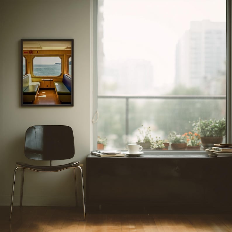May include: A framed wall art print depicting the interior of a ferry cabin with blue and white bench seating facing a window that looks out onto the ocean. The print is displayed on a light-colored wall next to a black chair and a window.