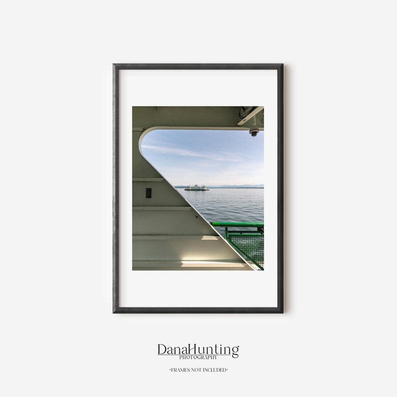 A vertical photograph of a Washington State Ferry boat gliding through Puget Sound, framed in the image by the unique angled architecture of another ferry boat.  Mountains and blue sky in background, green railing and ferry architecture in foreground